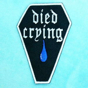 Op de afbeelding: Zwarte en witte geborduurde patch in de vorm van een kist met de tekst "died crying" in wit en een blauwe traan.