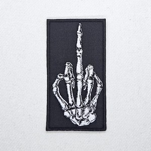 Pode incluir: Etiqueta bordada preta com uma mão de esqueleto branca mostrando o dedo do meio.