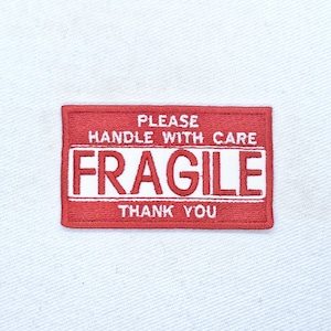 May include: A rectangular red patch with white embroidered text. The patch reads "PLEASE HANDLE WITH CARE FRAGILE THANK YOU". The patch is likely used for shipping or handling delicate items.