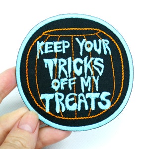 Może przedstawiać: Czarna, pomarańczowa i niebieska haftowana naszywka do prasowania z tekstem "Keep Your Tricks Off My Treats". Naszywka ma kształt dyni.