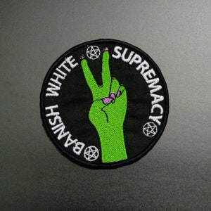 Puede incluir: Parche circular negro con texto bordado en blanco que dice "BANISH WHITE SUPREMACY". Una mano verde en señal de paz está en el centro, con uñas rosas. El parche tiene tres pentagramas blancos.