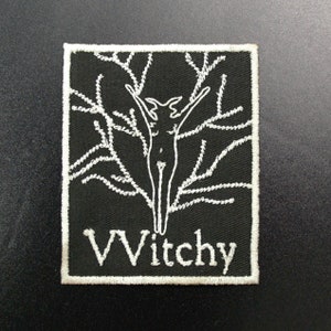 May include: Black embroidered patch with a white outline. The patch features a stylized figure with outstretched arms, surrounded by branches. The text "Witchy" is embroidered in white at the bottom.