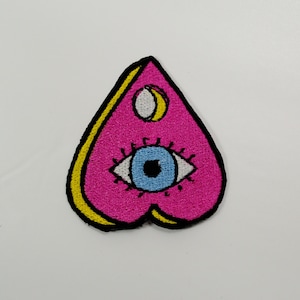 May include: A pink heart-shaped patch with a yellow outline and a black and white eye with a blue iris in the center. The eye is surrounded by black eyelashes. There is a white crescent moon above the eye.