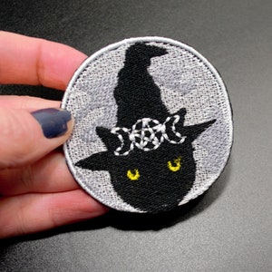 May include: A black cat wearing a witch's hat with a triple moon symbol embroidered on it. The cat is on a gray background that resembles a moon.