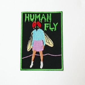 May include: A black and green embroidered patch with a human figure with a fly head. The text "HUMAN FLY" is embroidered in green on the patch.