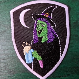 May include: Embroidered patch featuring a green-skinned witch with purple hair and a black hat, holding a blue mug. The witch is set against a black background with a crescent moon, framed by a white border. A fun, spooky accessory.
