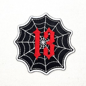 May include: A black and white spiderweb patch with the number "13" in red embroidered in the center.