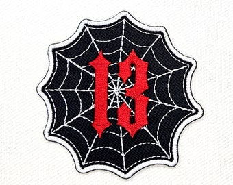 Op de afbeelding: Een zwarte en witte spinnenweb-patch met het nummer "13" in rood geborduurd in het midden.
