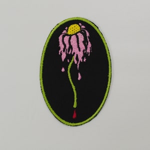 May include: A black oval embroidered patch with a green outline. The patch features a pink flower with a yellow centre, dripping with pink and red liquid.