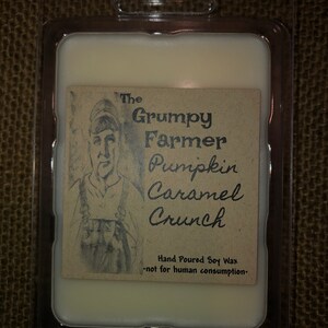 Puede incluir: Un ambientador de cera rectangular en un envase de plástico transparente. La etiqueta dice "The Grumpy Farmer Pumpkin Caramel Crunch" con un boceto de un granjero. También se ve el texto "Hand Poured Soy Wax" y "not for human consumption".