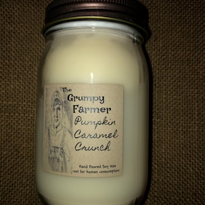 Puede incluir: Una vela en tarro con tapa marrón y una etiqueta que dice "The Grumpy Farmer Pumpkin Caramel Crunch". La etiqueta presenta un boceto de un granjero. La vela está rellena de cera cremosa blanquecina.