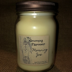 May include: A glass jar candle with a brown lid. The label reads "The Grumpy Farmer Morning Joe" and "Hand Poured Soy Wax - not for human consumption".