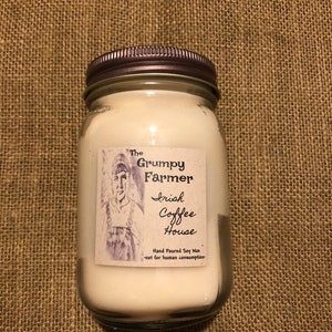 May include: A glass jar candle with a metal lid. The label reads "The Grumpy Farmer Irish Coffee House." The candle is filled with white wax and is hand-poured soy wax. The label also features a drawing of a person.