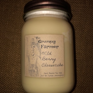 May include: A glass jar candle with a brown lid. The label reads "The Grumpy Farmer" and "Wild Berry Cheesecake". The label also states "Hand Poured Soy Wax" and "not for human consumption".