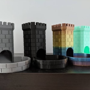 May include: Four 3D printed castle-shaped dice towers in different colors: gray, black, gold and green. Each tower has a brick pattern and a curved opening at the bottom for dice to roll out. The towers are sitting on a wooden surface.