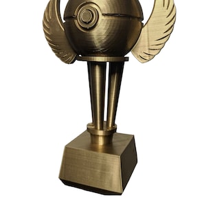 May include: A gold-colored trophy with a round, winged base and a sphere on top. The sphere has a circular design on it.
