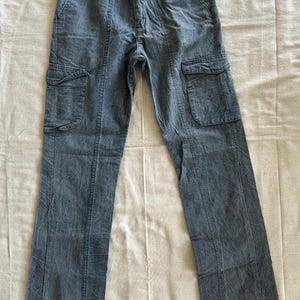 May include: A pair of blue-grey cargo pants with two large pockets on each leg. The pants have a button and zipper closure and are made of a textured fabric. The pants are laid flat on a neutral-colored surface.