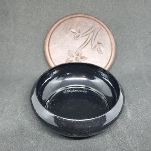 May include: A black ceramic dish with a shiny finish and a round, slightly raised rim. The dish is empty and has a small, circular indentation in the center. The dish is sitting on a gray surface.