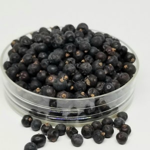 May include: A clear glass dish filled with dark blue juniper berries. Some berries are scattered around the dish on a white surface. The berries have a slightly wrinkled texture.
