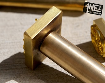 May include: Close-up of a brass stamp with a square head and a round handle. The stamp has a textured surface and a small, raised design on the head. The handle is smooth and polished.