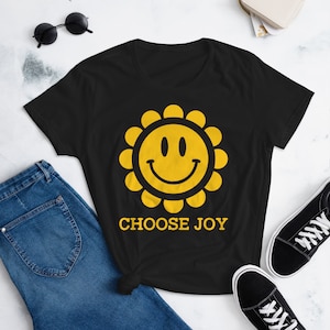 May include: Black t-shirt featuring a yellow smiley face design with a sunflower outline. The text "CHOOSE JOY" is printed below the graphic. The shirt is paired with blue jeans and black sneakers.