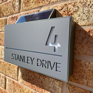 May include: A gray metal house number sign with a solar panel on top. The sign reads "4 STANLEY DRIVE".