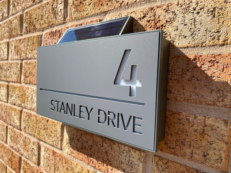 May include: A gray metal house number sign with a solar panel on top. The sign reads "4 STANLEY DRIVE".
