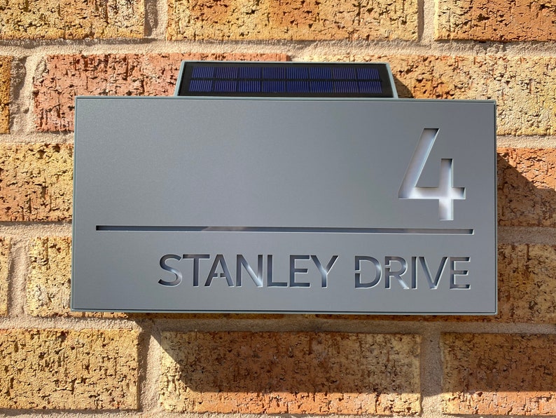 May include: A gray house number sign with a solar panel on top. The sign has the number "4" and the street name "STANLEY DRIVE" cut out.