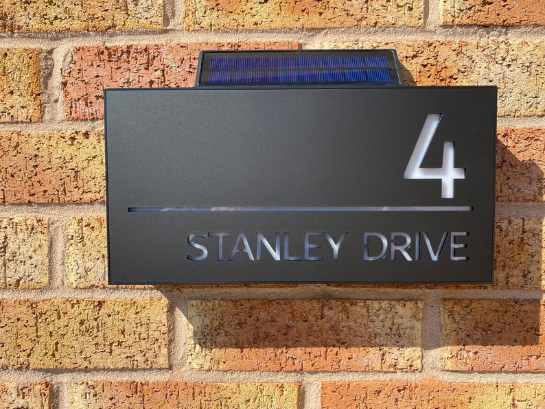 May include: A black metal house number sign with a solar panel on top. The sign has white cut-out lettering that reads "4 STANLEY DRIVE".
