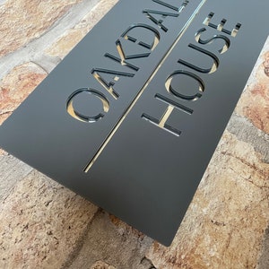 May include: A dark gray rectangular sign with the words "Oakdale House" cut out in a white font.