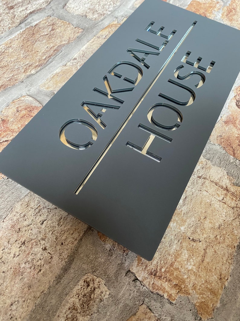 May include: A dark gray rectangular sign with the words "Oakdale House" cut out in a white font.