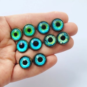 May include: Assortment of ten blue and green glass eyes with black pupils, set in bronze settings. The eyes are arranged in a semi-circle on a hand.