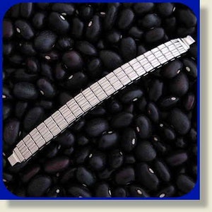 May include: A silver expandable watch band with a polished finish. The band is laid out on a bed of black beans.