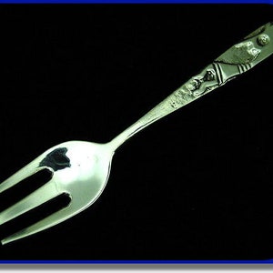 May include: A silver fork with a decorative handle featuring a stylised figure. The fork has three tines and a rounded handle.