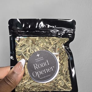 May include: A black resealable pouch containing a blend of dried green herbs and botanical pieces. A circular label on the front features a white star icon and the text: Made Divinely Conjure Shop, Road Opener, Made with Intention.