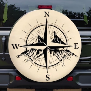 May include: A beige spare tire cover featuring a black compass rose design with mountain silhouettes. The compass points North, South, East, and West. The cover is mounted on the back of a black Jeep.