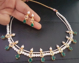 May include: A double-strand pearl necklace with green gemstone accents. The necklace has a gold chain and is adorned with small, square-shaped pendants with clear stones. The necklace is paired with matching earrings.