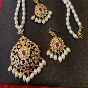 May include: A pearl necklace with a gold pendant and earrings. The pendant and earrings feature a circular design with a pink stone in the center and smaller stones around it.