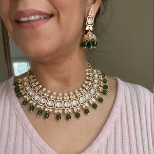 May include: A gold necklace with a green and white design. The necklace features a row of square-shaped stones with smaller stones around them. The necklace is accented with green beads.