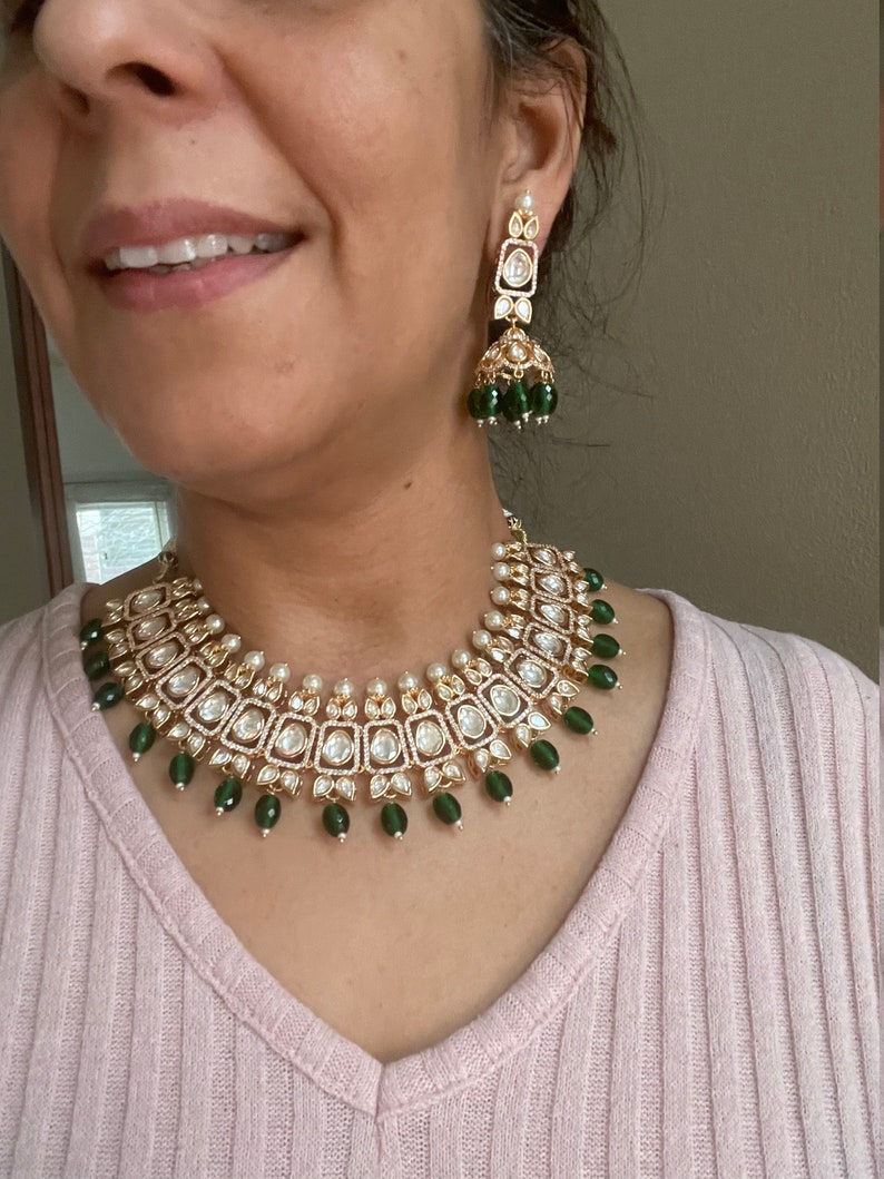 May include: A gold necklace with a green and white design. The necklace features a row of square-shaped stones with smaller stones around them. The necklace is accented with green beads.