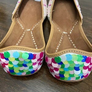May include: A pair of white flat shoes with colorful sequins on the toes. The shoes have a brown leather insole and a white fabric upper.