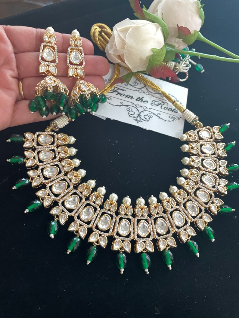 May include: A gold and white necklace with green accents. The necklace features a square design with multiple layers of stones. The necklace is paired with matching earrings.