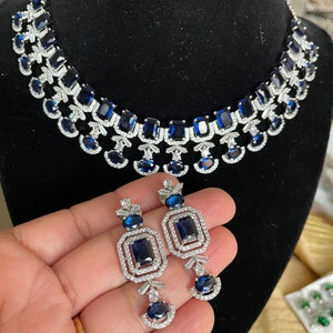 May include: A silver necklace and earrings set with blue gemstone accents. The necklace features a layered design with multiple rows of blue stones and clear rhinestones. The earrings are drop style with a rectangular blue stone surrounded by clear rhinestones.
