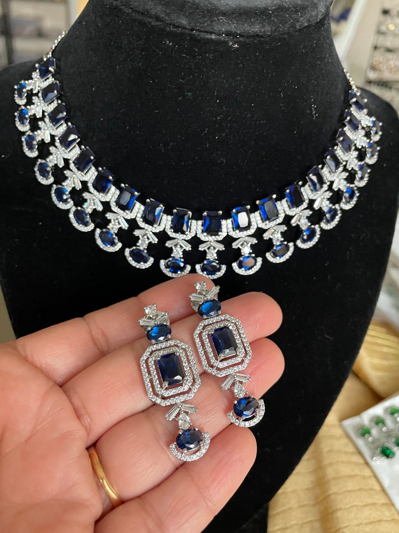 May include: A silver necklace and earrings set with blue gemstone accents. The necklace features a layered design with multiple rows of blue stones and clear rhinestones. The earrings are drop style with a rectangular blue stone surrounded by clear rhinestones.