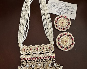 May include: A white pearl necklace with a gold pendant featuring a red and white stone design. The pendant has a long, dangling section with multiple layers of pearls and red stones. The necklace is paired with matching earrings with a similar design.