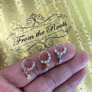 May include: Three nose rings with a dangling diamond charm. Each ring is made of a different metal: gold, silver, and rose gold. The rings are set with clear cubic zirconia stones.