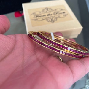 May include: Two gold bangles with a row of pink square stones set in the metal. The bangles are stacked on top of each other.