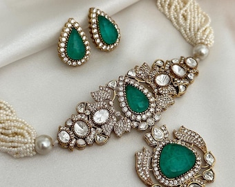Op de afbeelding: Een sierlijke sieradenset met een ketting en bijpassende oorbellen. De ketting heeft een goudkleurige metalen basis met smaragdgroene druppelvormige stenen en heldere kristallen. De oorbellen weerspiegelen het druppelontwerp. De ketting is geregen met witte kralen.