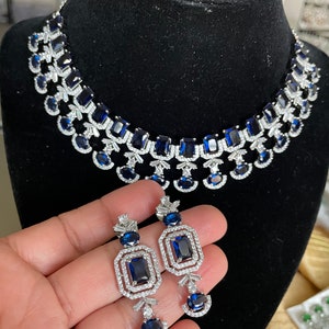 May include: A silver necklace and earrings set with blue gemstones. The necklace features a double row of rectangular and round stones, while the earrings have a single rectangular stone surrounded by smaller stones.