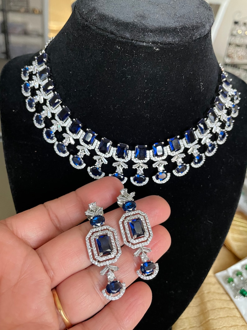 May include: A silver necklace and earrings set with blue gemstones. The necklace features a double row of rectangular and round stones, while the earrings have a single rectangular stone surrounded by smaller stones.